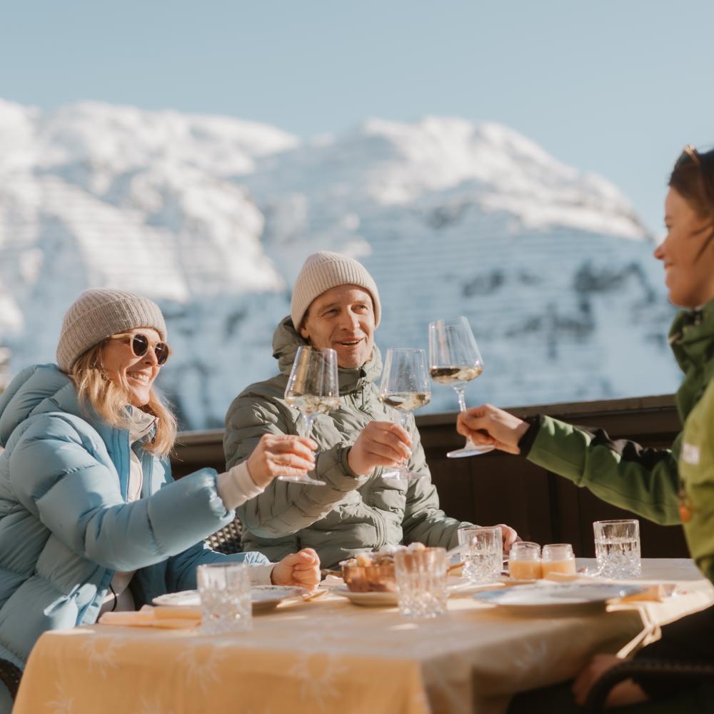 Gäste stoßen mit Getränken auf der Terrasse des Hotel Post Lech mit verschneiten Bergen im Hintergrund an