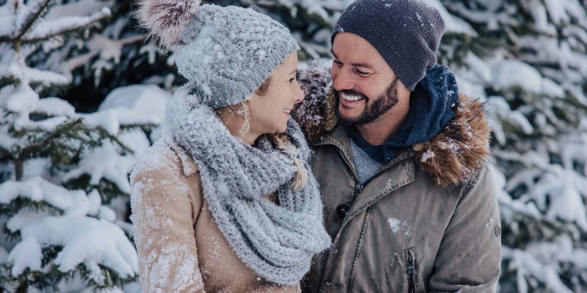 A happy couple are enjoying their winter holiday at the Designer Motel Glockna in Fusch am Großglockner, set against a snow-covered forest backdrop.