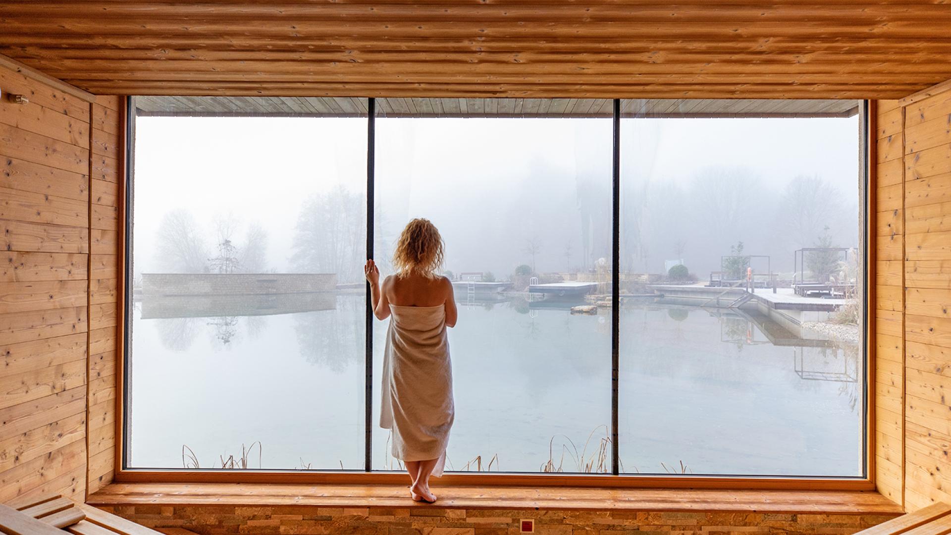 Gast im Holz-Spa des Pfalzblick Wald Spa Resort blickt durch ein Panoramafenster auf neblige Außenbecken