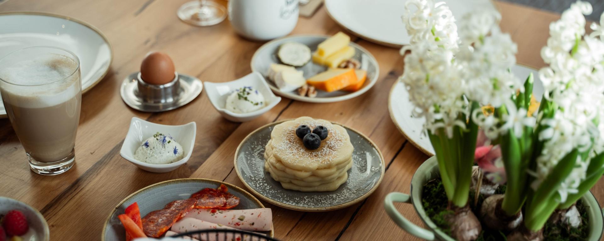 Frühstückstisch mit Ei, Käse, Aufstrich, Wurst, Kaffee, Pancakes und einem Glas Orangensaft