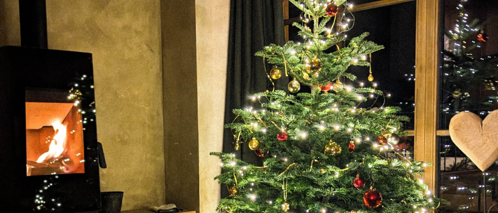 eihnachtlich geschmückter Christbaum mit Lichterkette und roten und goldenen Kugeln im Wohnraum eines Chalets. Links brennt Feuer im Kaminofen mit Holzlege darunter, rechts steht ein großes Holzherz vor dem bodentiefen Fenster.