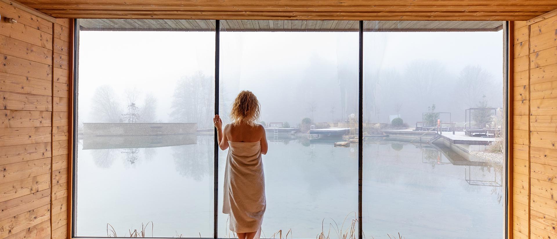 Gast im Holz-Spa des Pfalzblick Wald Spa Resort blickt durch ein Panoramafenster auf neblige Außenbecken