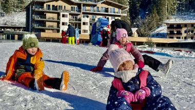 Kinder rodeln im Schnee vor dem Familienresort Buchau in winterlicher Waldlandschaft