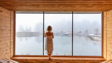 Gast im Holz-Spa des Pfalzblick Wald Spa Resort blickt durch ein Panoramafenster auf neblige Außenbecken