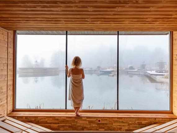 Gast im Holz-Spa des Pfalzblick Wald Spa Resort blickt durch ein Panoramafenster auf neblige Außenbecken
