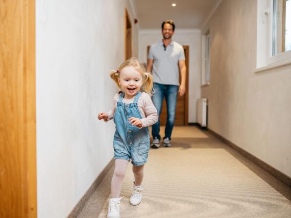 Fröhliches Kind läuft im Flur des Familienresort Buchau, Vater im Hintergrund