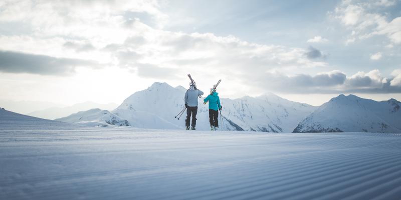 Zwei Skifahrer mit Skiern bei Hotel Seeber vor schneebedeckten Bergen