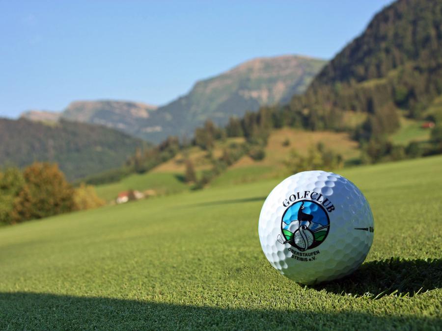 Golfball auf grünem Rasen mit Allgäuer Bergen und Sonne im Hintergrund